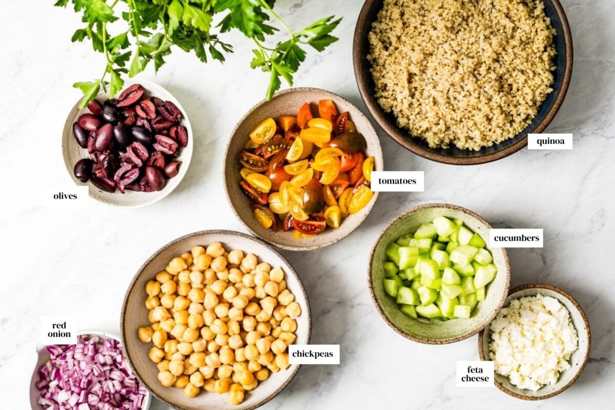 Labeled ingredients for Mediterranean quinoa salad, including quinoa, chickpeas, cherry tomatoes, cucumber, red onion, Kalamata olives, and feta cheese.