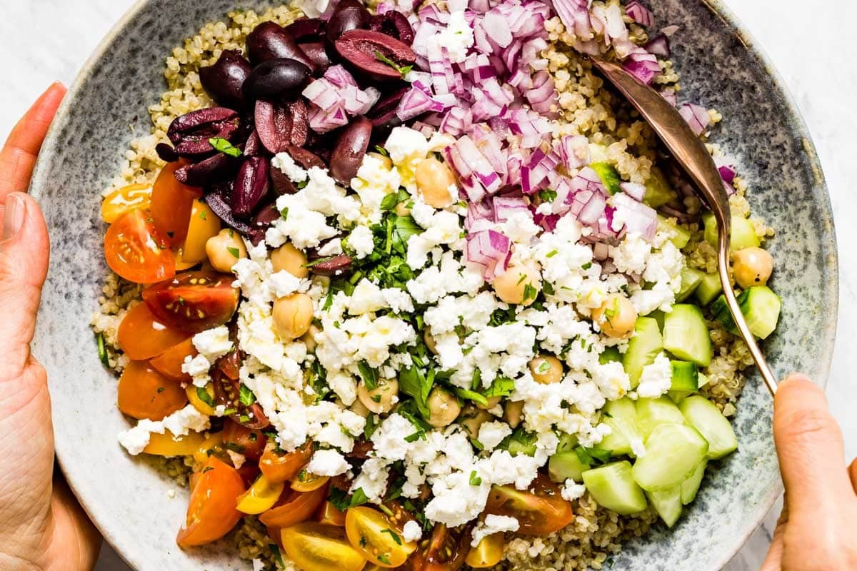 Hands holding a bowl of quinoa topped with sectioned piles of feta, Kalamata olives, red onion, and cucumber before tossing.