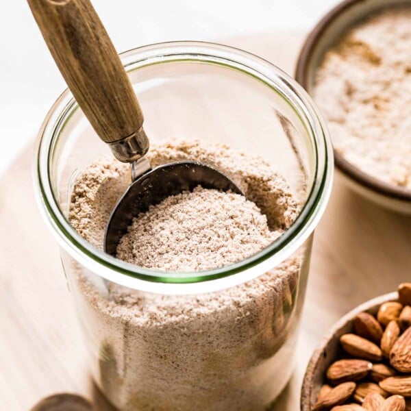 A jar of almond pulp with whole raw almonds on the side in a bowl