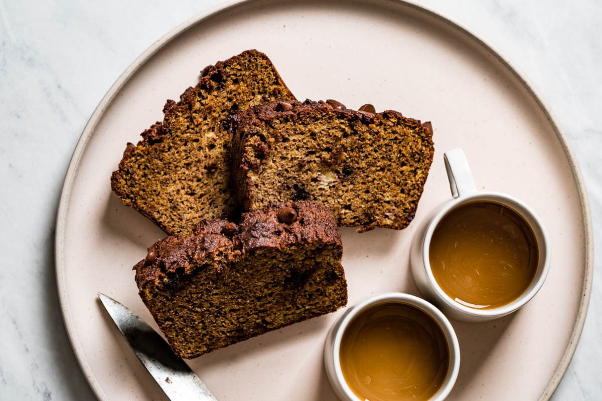 Sliced almond banana bread served on a plate with two cups of coffee.