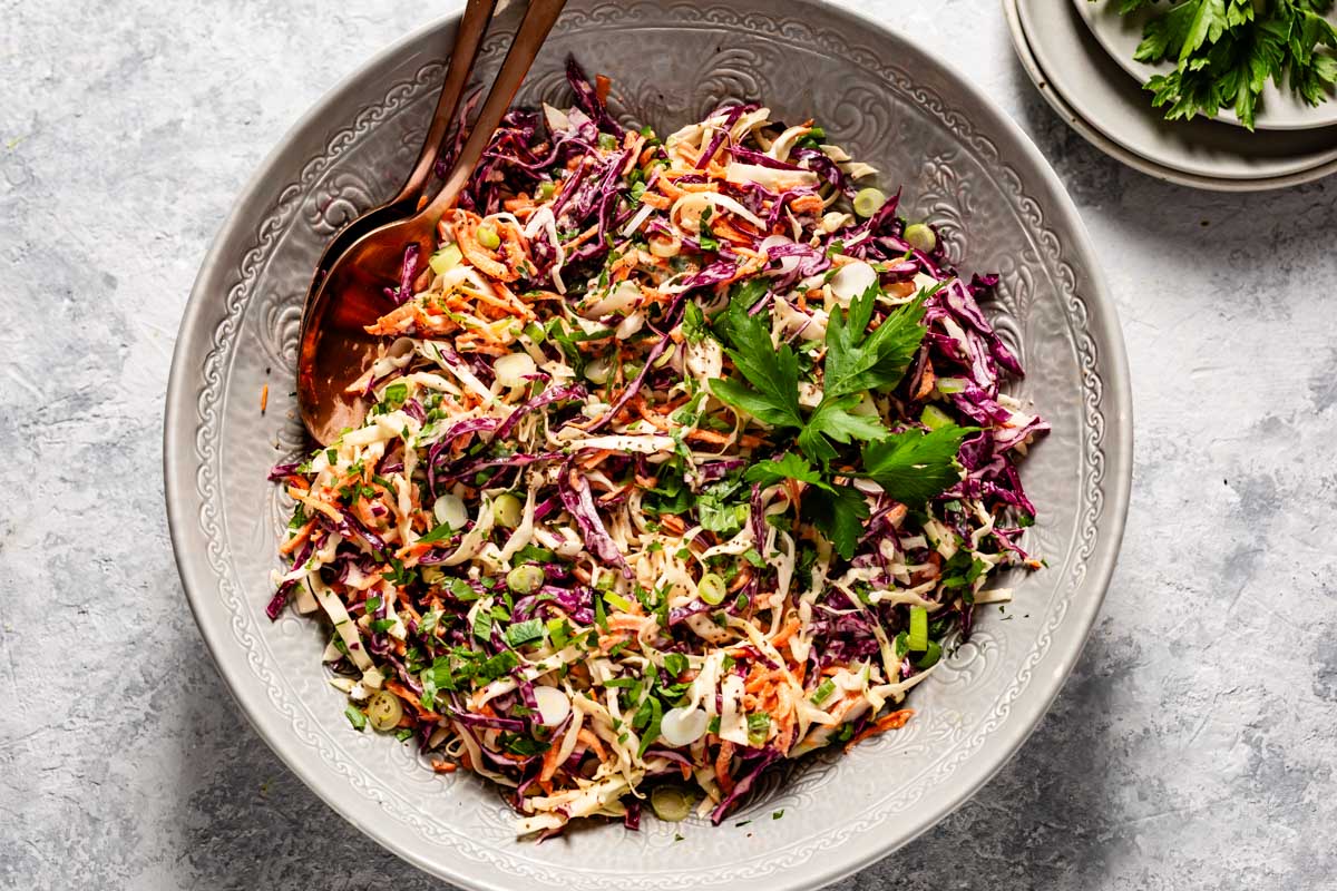 Cabbage salad with Greek yogurt dressing tossed with carrots, green onions, and parsley, served in a grey bowl with a serving spoon.