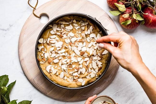 Paleo apple cake batter in a pan being sprinkled with sliced almonds by a person top view
