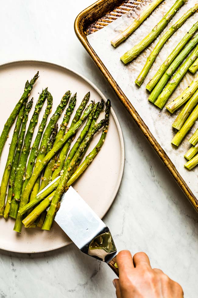 Baked asparagus being transferred from baking sheet to a plate by a person