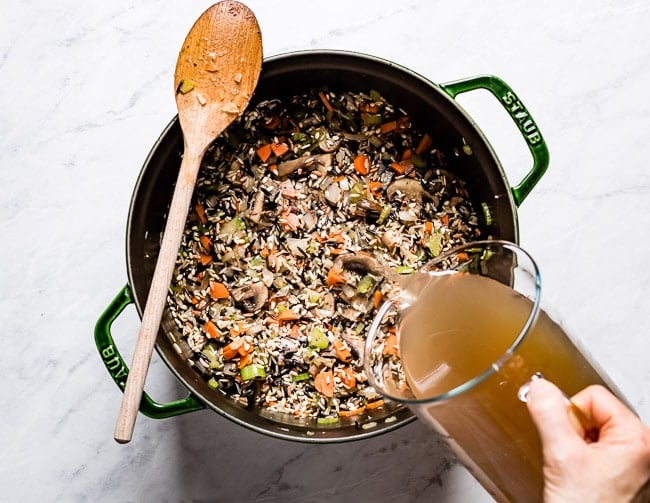 Person pouring stock into a large pot with rice and veggies