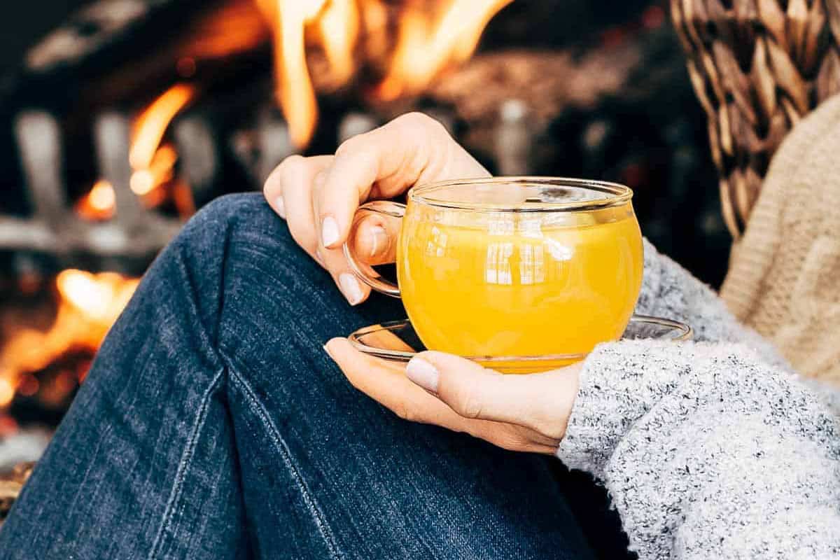 A cozy lifestyle image of a person sitting by a warm fireplace holding a steaming mug of vibrant ginger turmeric tea.