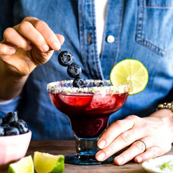 Hands placing a blueberry garnish into a salted margarita glass filled with a dark purple blueberry margarita and ice.