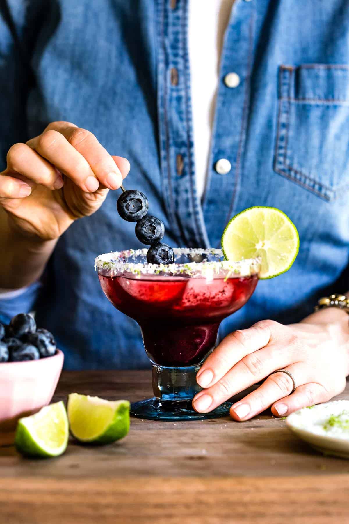 Hands placing a blueberry garnish into a salted margarita glass filled with a dark purple blueberry margarita and ice.