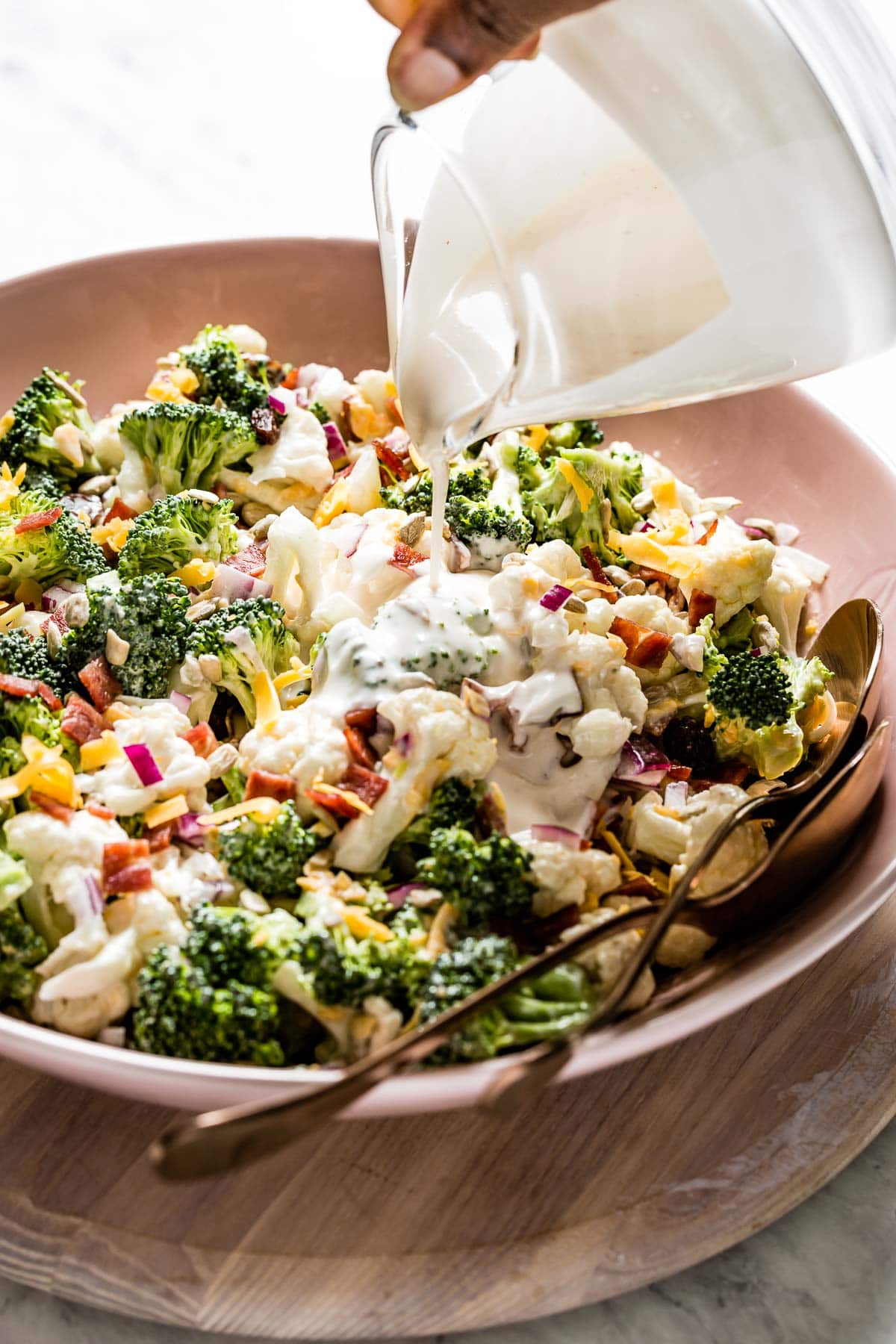 Person drizzling broccoli cauliflower salad with bacon with dressing.