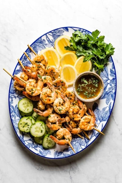 Grilled shrimp skewers on a plate with lemon, herbs and cucumber slices