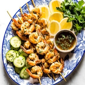 grilled shrimp on skewers on a plate with lemon slices, herbs, and cucumber slices
