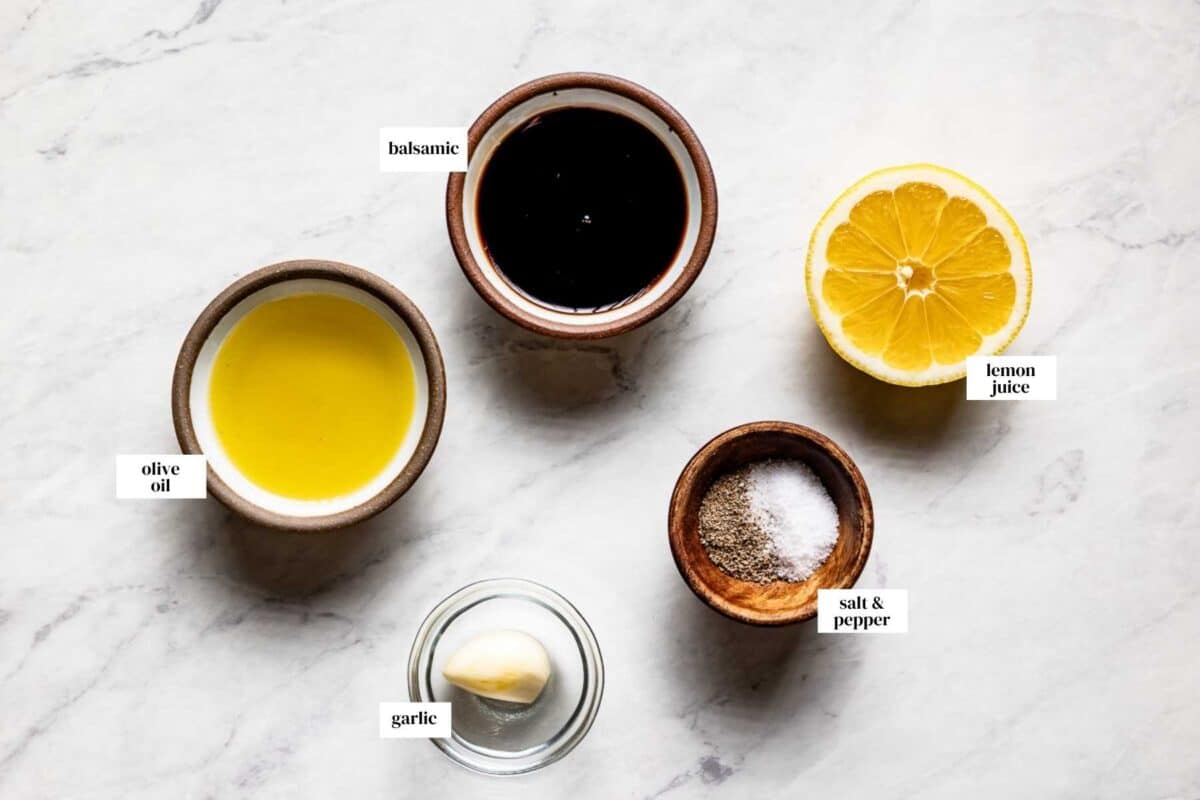 Labeled ingredients for balsamic lemon dressing including balsamic vinegar, fresh lemon, olive oil, garlic, salt, and pepper arranged on a marble surface.