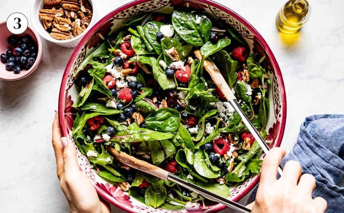 Hands tossing a spinach blueberry salad with feta, raspberries, and toasted pecans in a large patterned bowl using tongs.