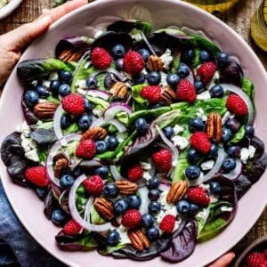 Overhead close-up of a blueberry salad with spinach, raspberries, crumbled feta, toasted pecans, and sliced red onion in a pink bowl, with a hand visible at the edge of the frame.