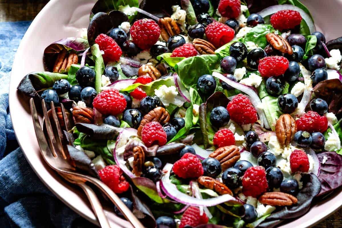Close-up of a spinach blueberry salad with crumbled feta, raspberries, toasted pecans, and sliced red onion in a pink bowl with a fork resting on the side.