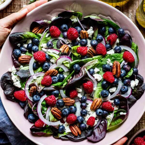 Hands holding a large bowl filled with blueberry spinach salad topped with raspberries, crumbled feta, toasted pecans, and sliced red onion, shot from above on a wooden surface.