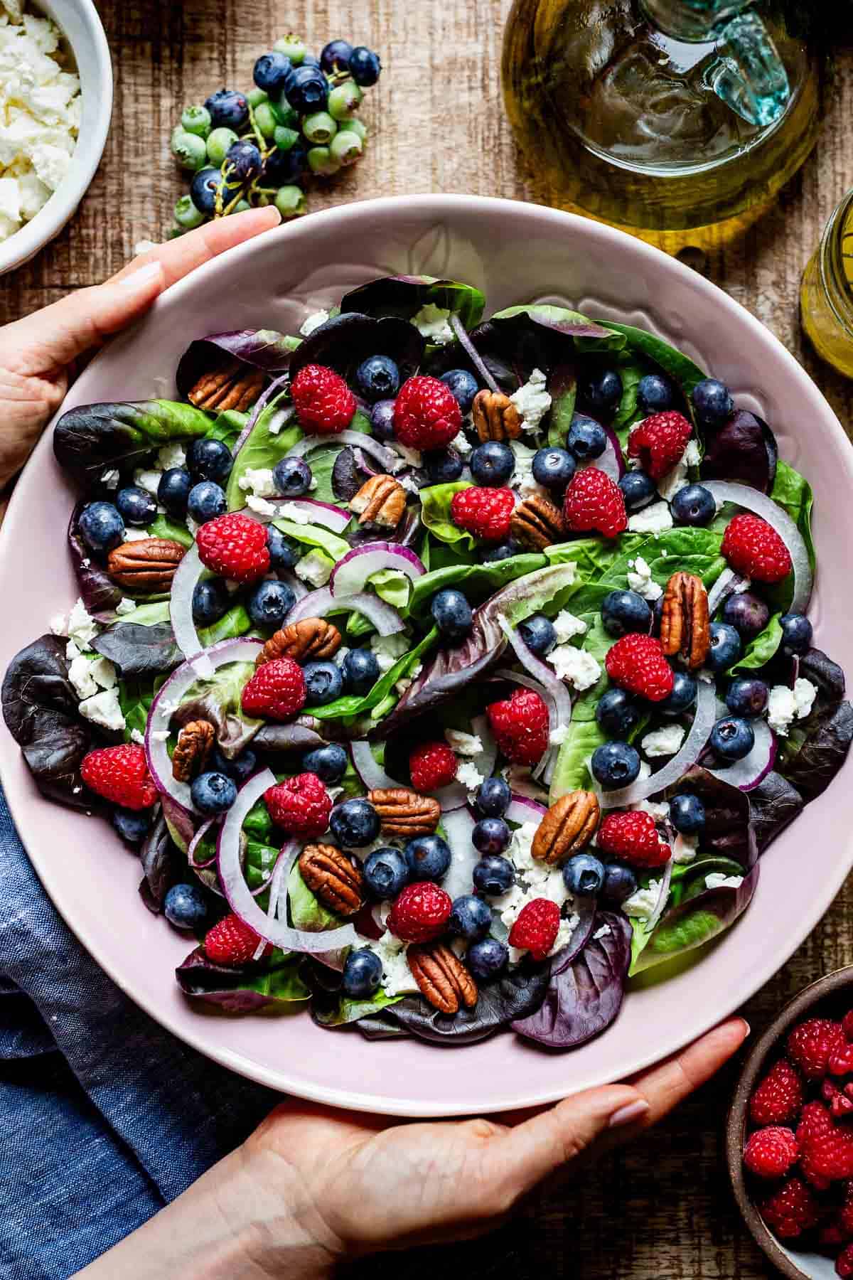 Hands holding a large bowl filled with blueberry spinach salad topped with raspberries, crumbled feta, toasted pecans, and sliced red onion, shot from above on a wooden surface.