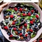 Overhead shot of a blueberry spinach salad with raspberries, pecans, red onion, and feta cheese in a pink bowl, held by two hands, with text overlay reading Blueberry Spinach Salad with Balsamic Dressing.