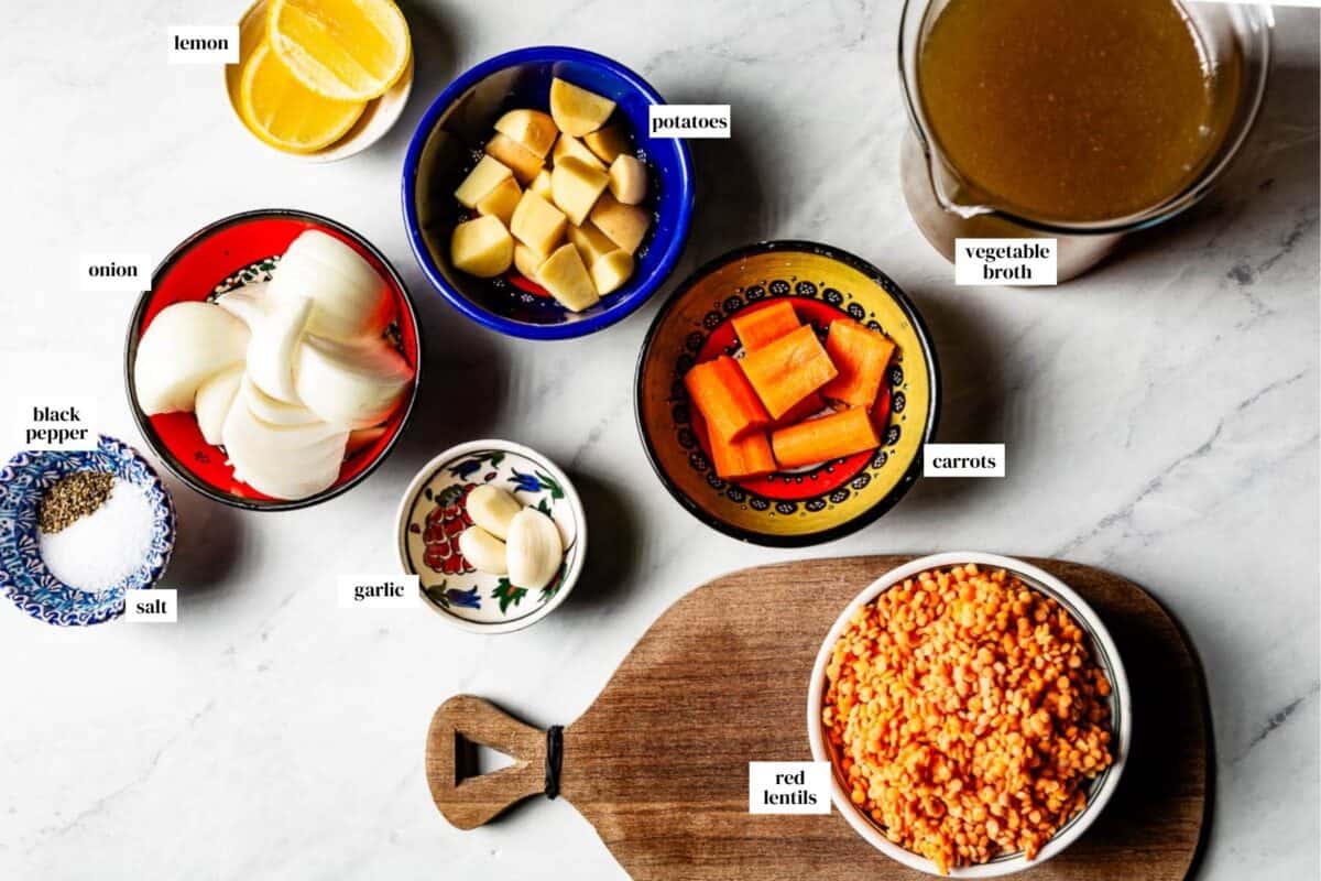 Ingredients for Turkish red lentil soup organized in small colorful bowls on a marble countertop, including red lentils, chopped onions, potatoes, carrots, garlic, vegetable broth, and lemon slices.