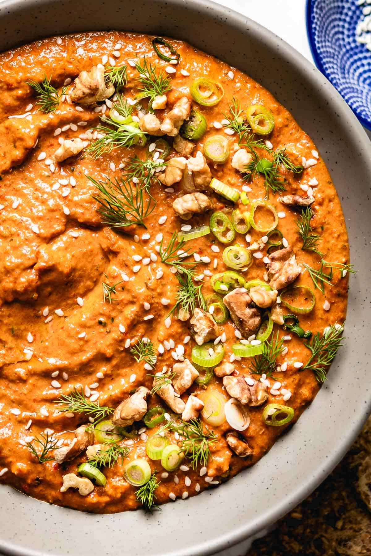 Muhammara is served in a large bowl and garnished with walnuts, scallions, and dill. 
