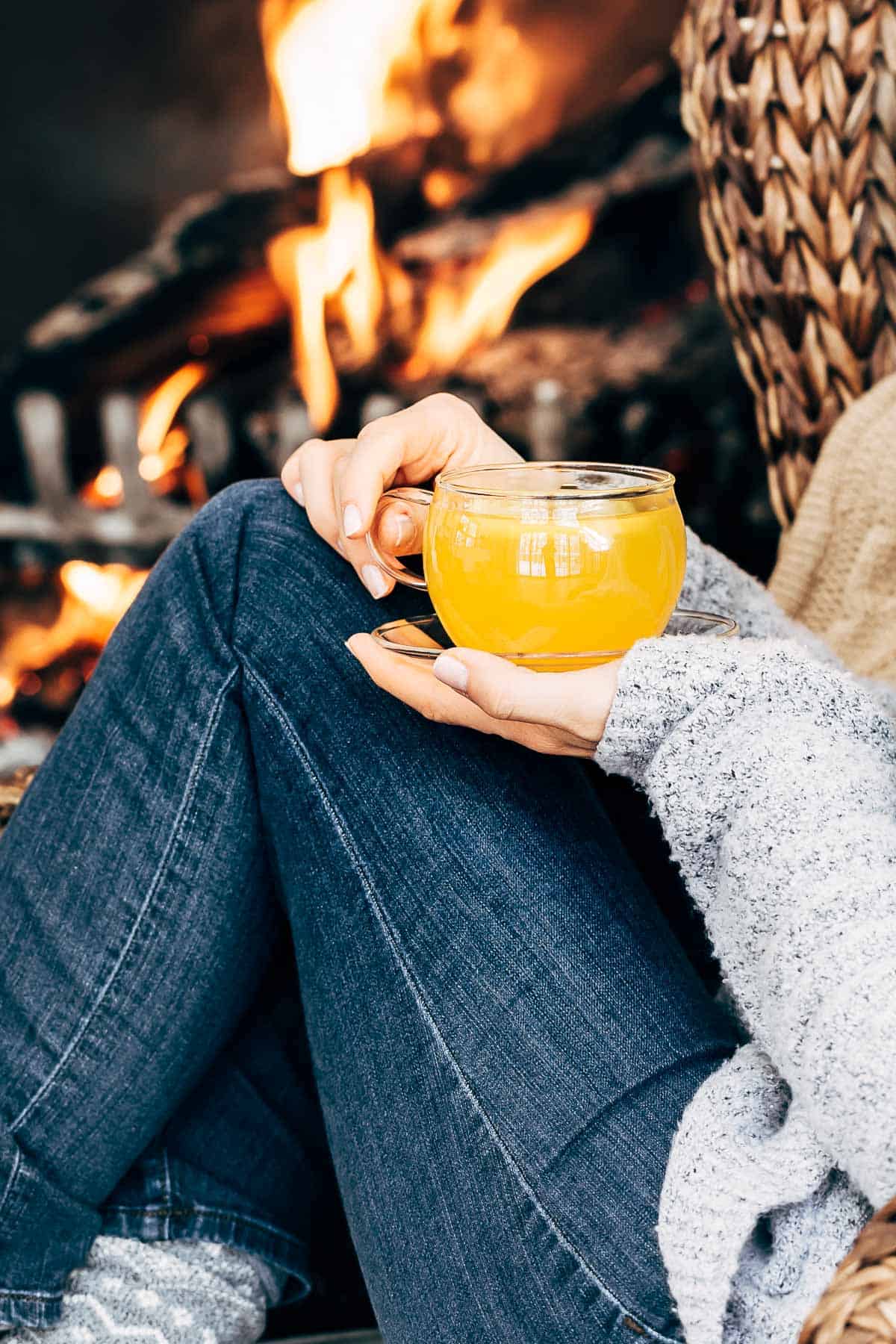 A woman is photographed from the front view as she is holding a glass of turmeric ginger tea behind a fire place.