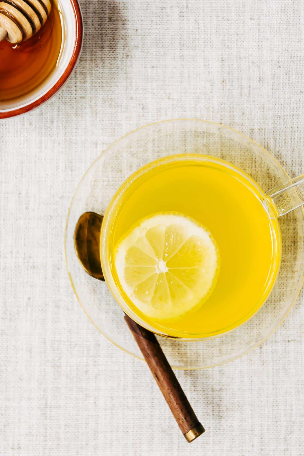 A glass cup of turmeric ginger tea with a lemon slice is photographed from the top view with a bowl of honey and sliced lemons.