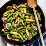 sauteed asparagus and mushrooms in a white skillet with a wooden spoon on the side
