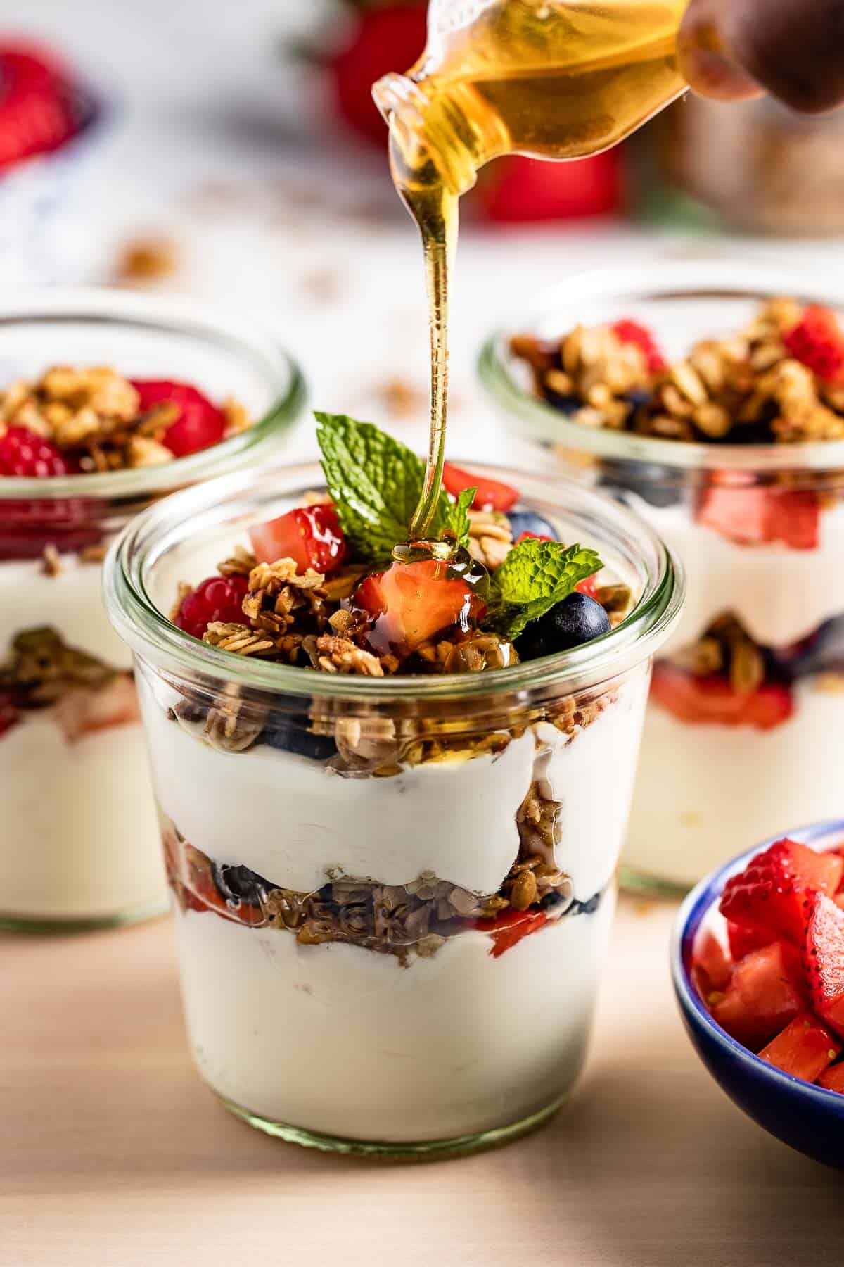 person drizzling strawberry blueberry granola parfait with honey