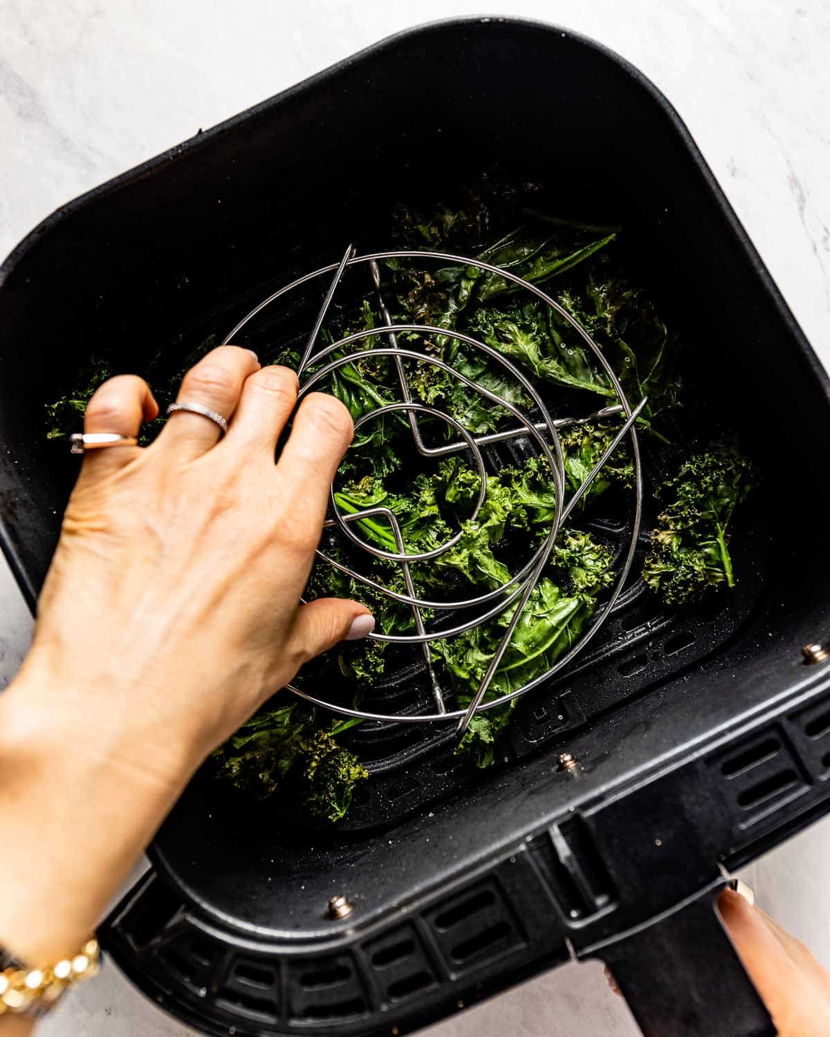 A person placing a rack over pieces of kale in an air fryer.