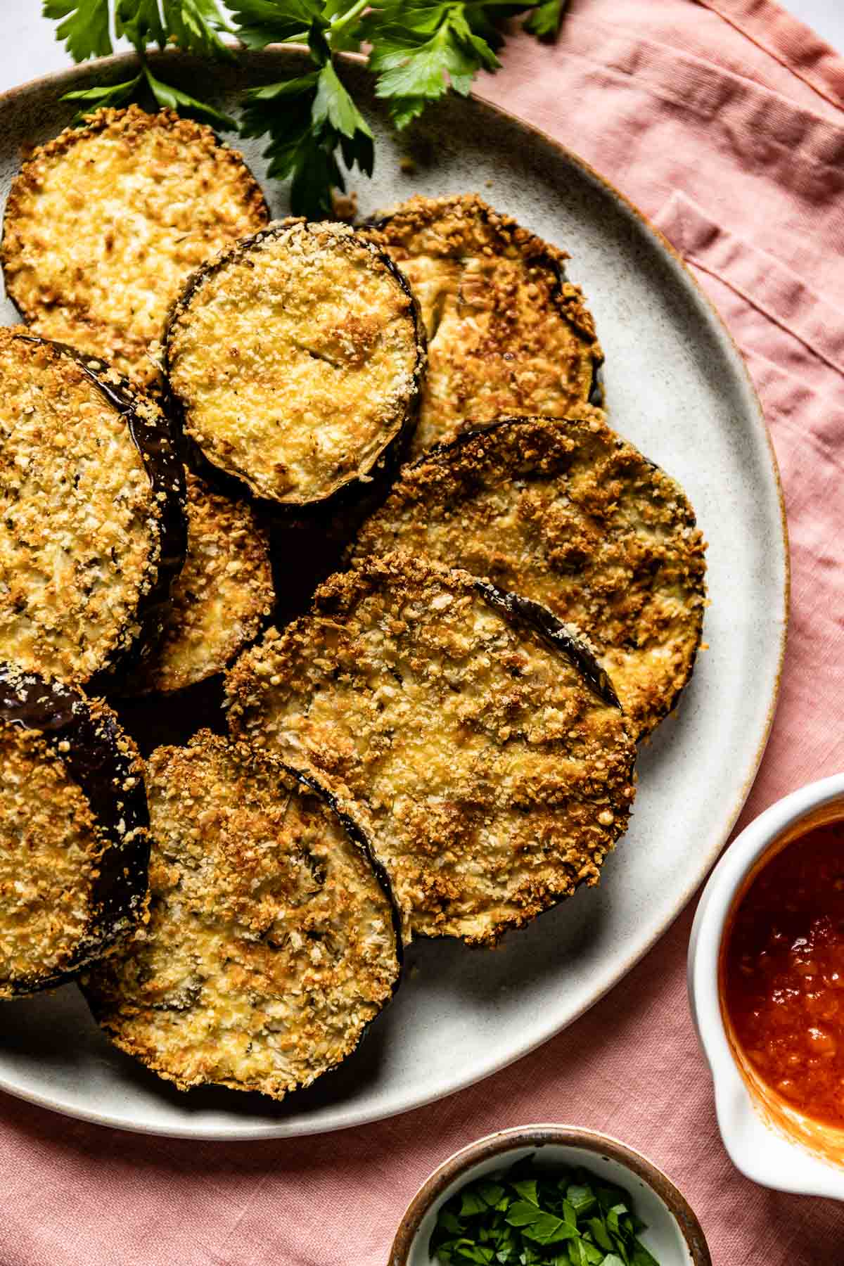 Air fried breaded eggplant on a plate with marinara sauce and fresh parsley on the side.