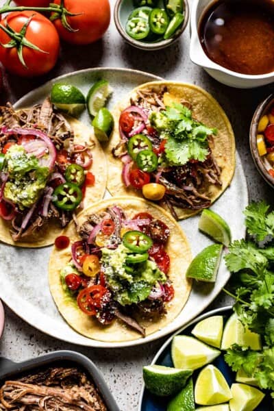 Brisket Tacos served over corn tortillas on a plate with toppings like guacamole, jalapeno, and tomatoes.