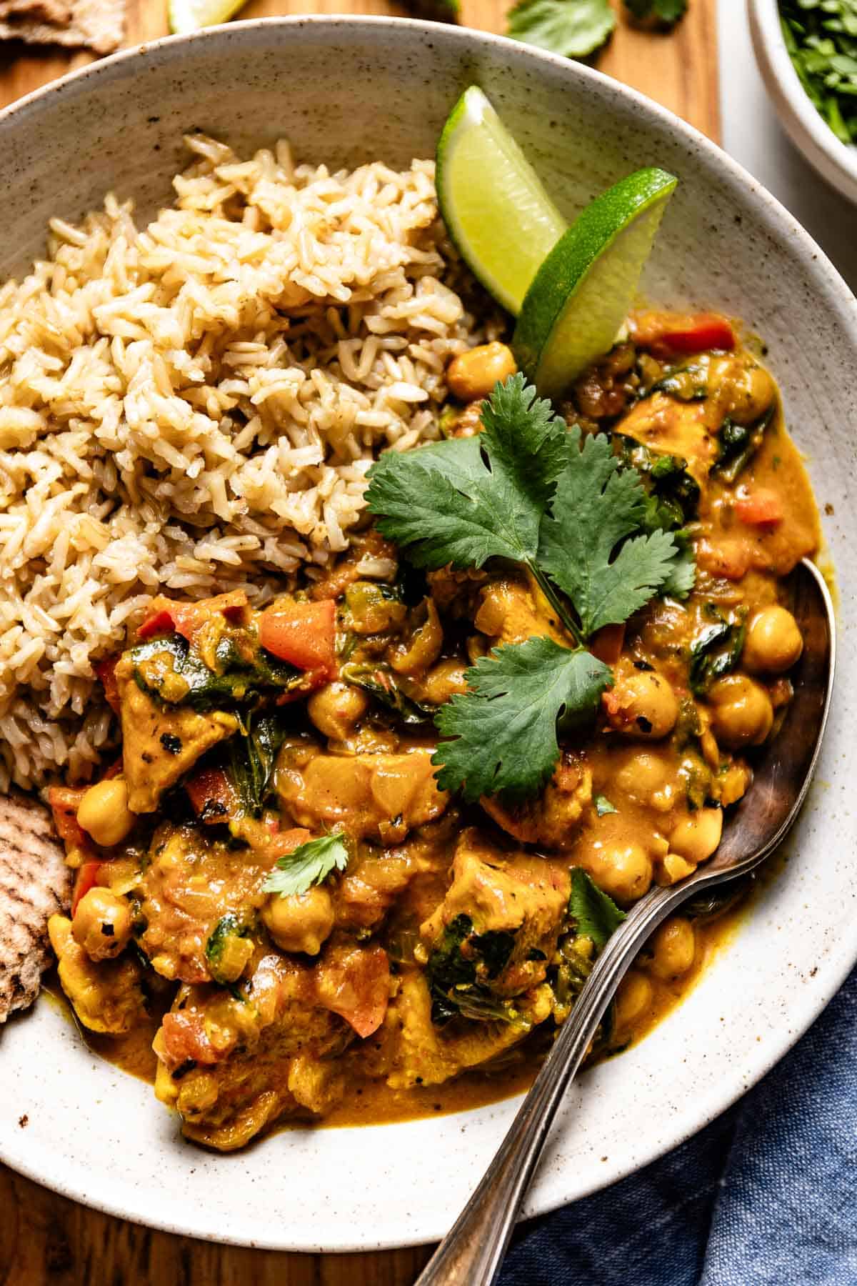 Chicken and chickpea curry in a bowl served with rice and lime wedges on the side.