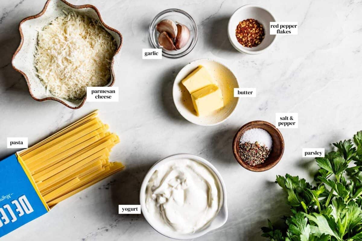 Flat lay of ingredients for Greek yogurt pasta sauce including fettuccine, a bowl of yogurt, garlic cloves, and fresh parsley on a marble surface.