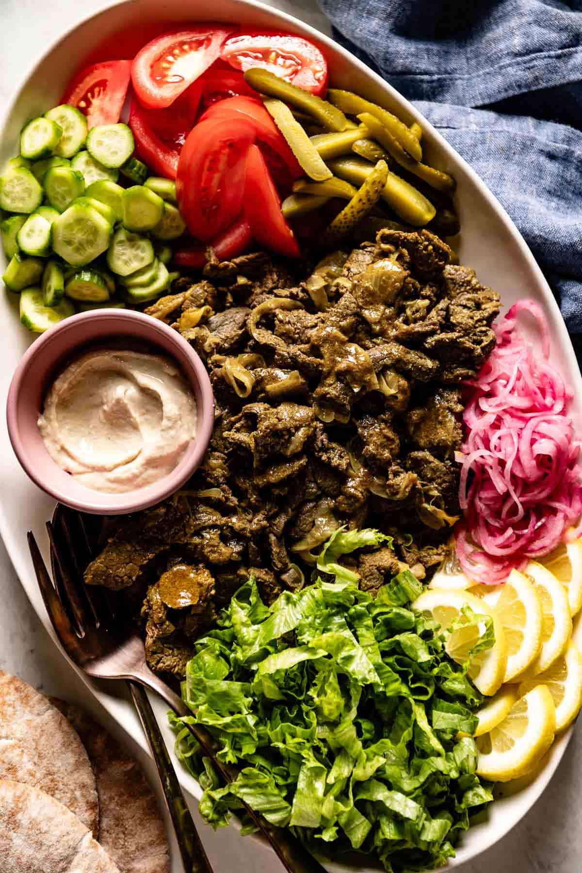 Steak shawarma on a plate with pickles, tahini sauce, tomatoes and salad greens.