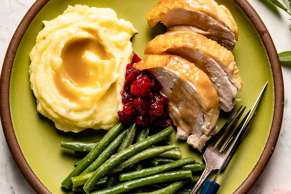 Roasted turkey breast served with gravy, mashed potatoes, green beans and cranberry sauce on a plate.