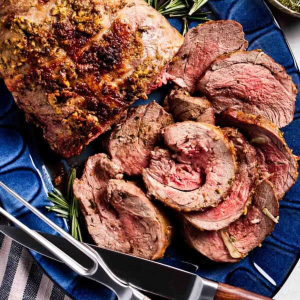 Roasted boneless leg of lamb on a plate sliced with utensils on the side.