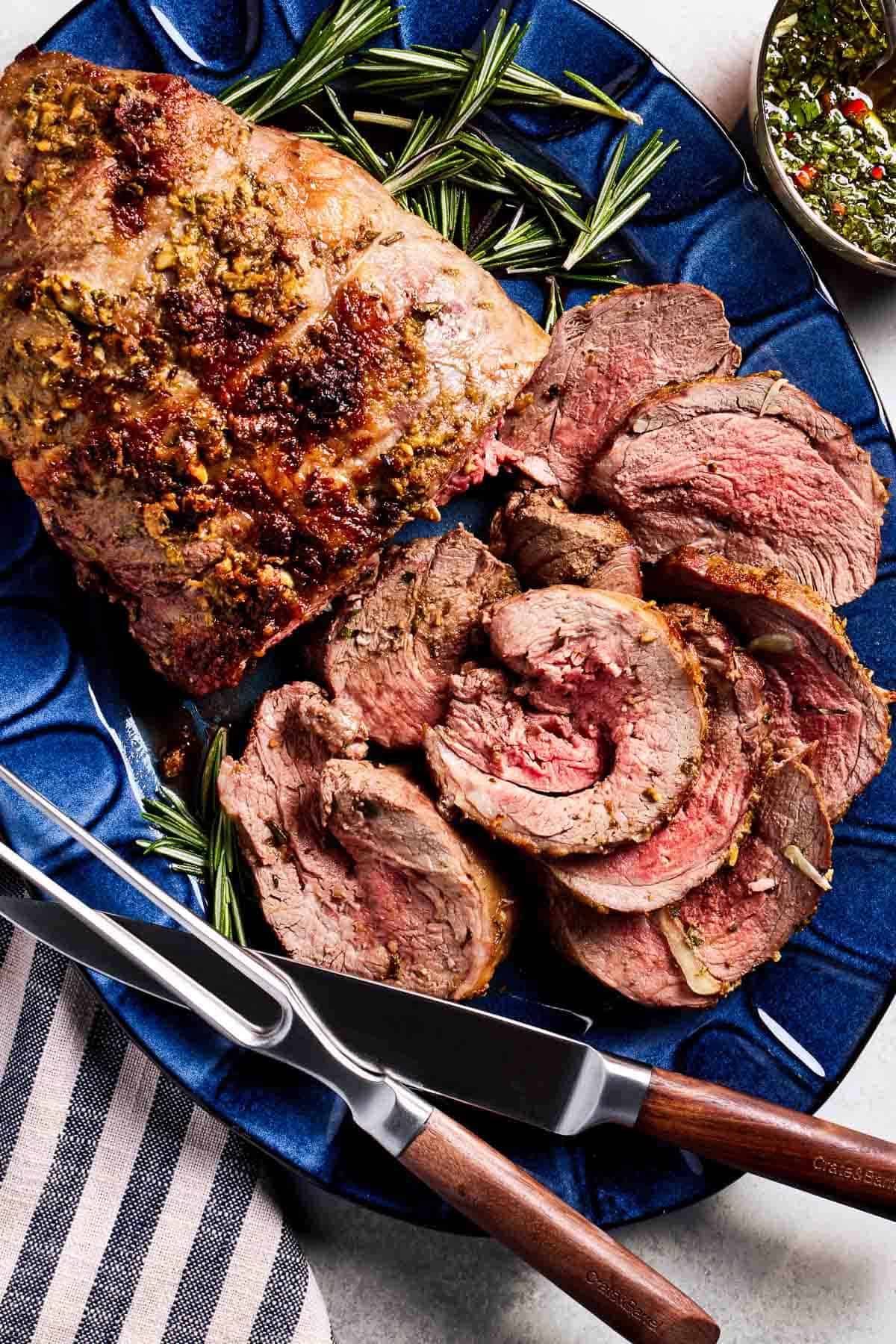 Roasted boneless leg of lamb on a plate sliced with utensils on the side.