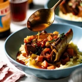 A close-up of a short rib being drizzled with savory pan sauce, served in a blue bowl over mashed vegetables.