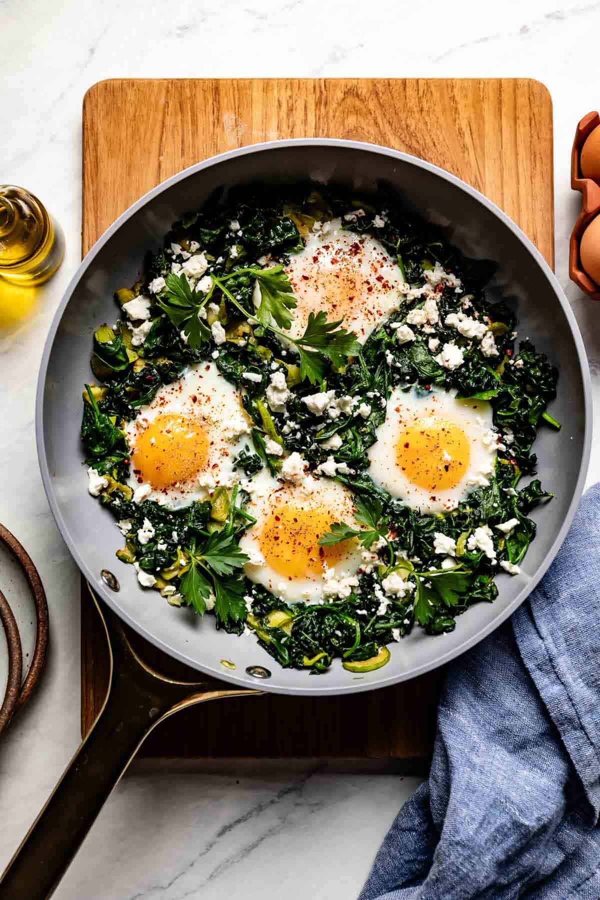 Top view of Green Shakshuka in a grey skillet with four runny eggs, kale, spinach, and feta.