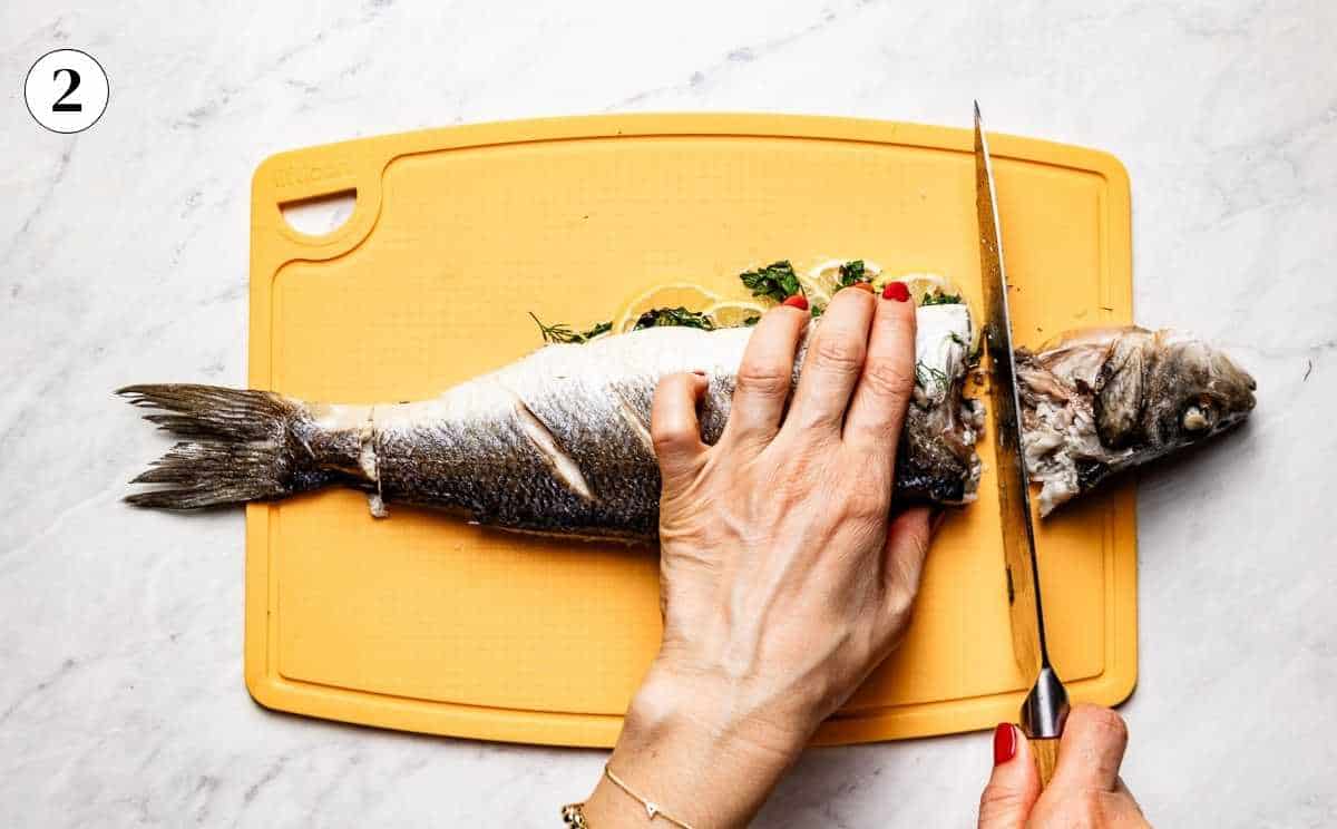 Removing the head of a roasted branzino with a sharp knife on a yellow cutting board.