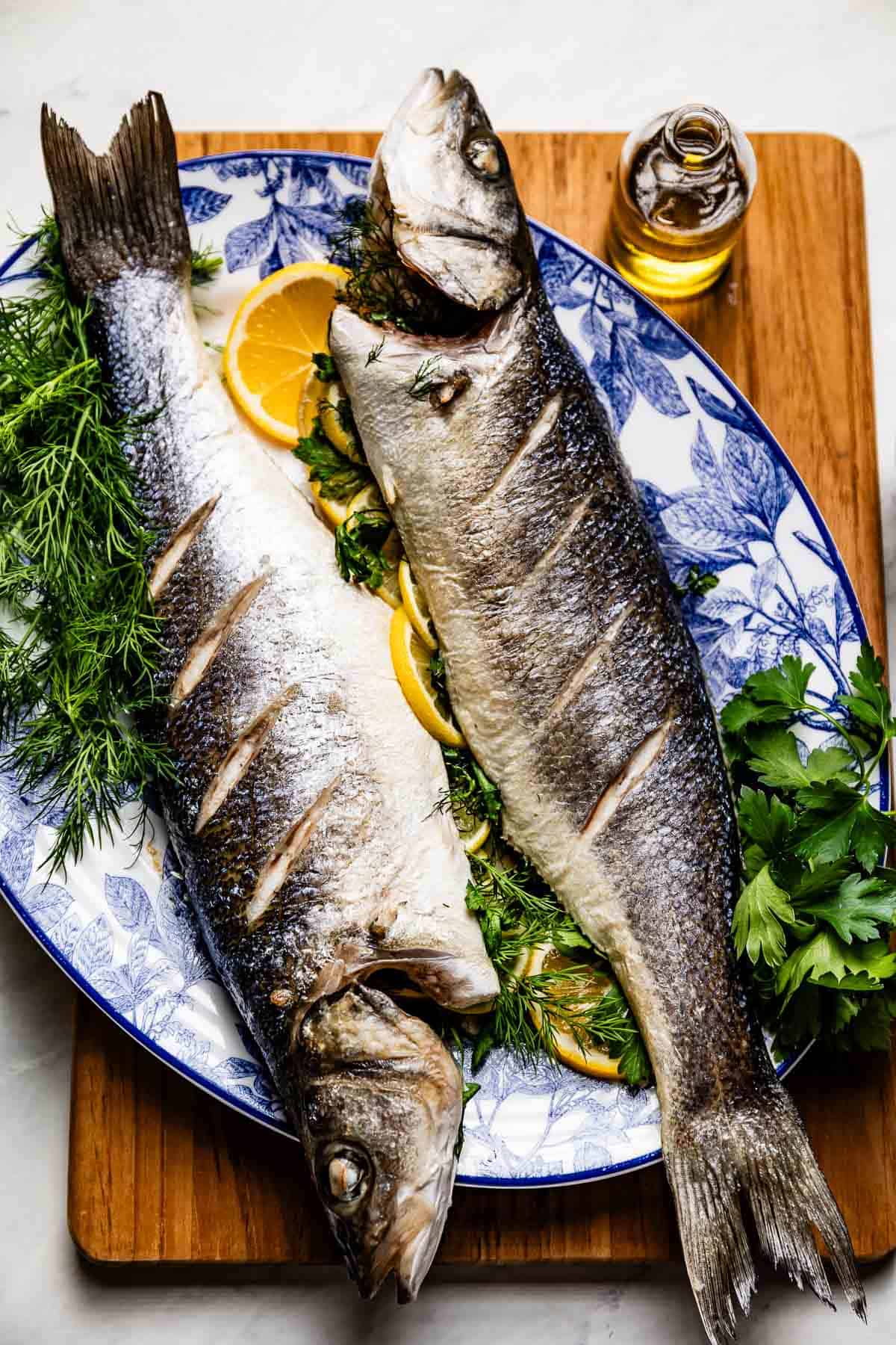 Two whole roasted Mediterranean sea bass on a blue and white plate garnished with lemon slices and fresh herbs.