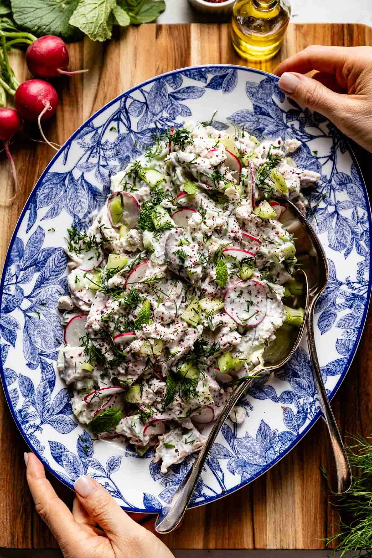 A portrait shot of a decorative blue platter filled with no-mayo chicken salad, held by hands at the edges, garnished with fresh dill and sumac.
