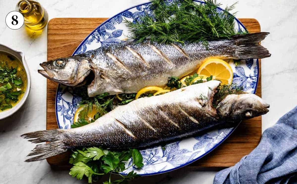 Two whole roasted branzino on a Mediterranean platter with fresh herbs, lemon, and olive oil sauce.