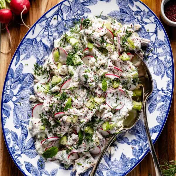 A portrait view of the final Tzatziki Chicken Salad on a decorative blue and white patterned platter with peeled diced cucumbers, radishes, feta, fresh dill, and sumac, on a wooden board.