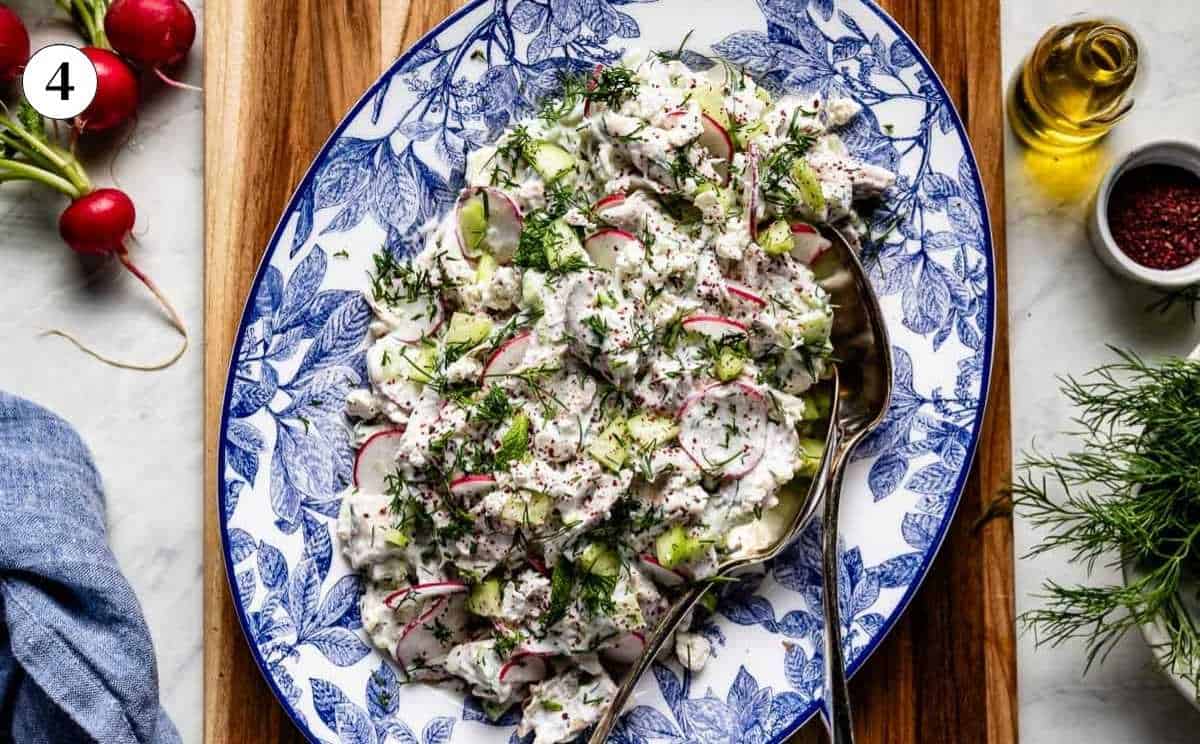 A horizontal view of creamy Tzatziki Chicken Salad with radishes and peeled cucumbers served on a blue and white platter on a wooden board.