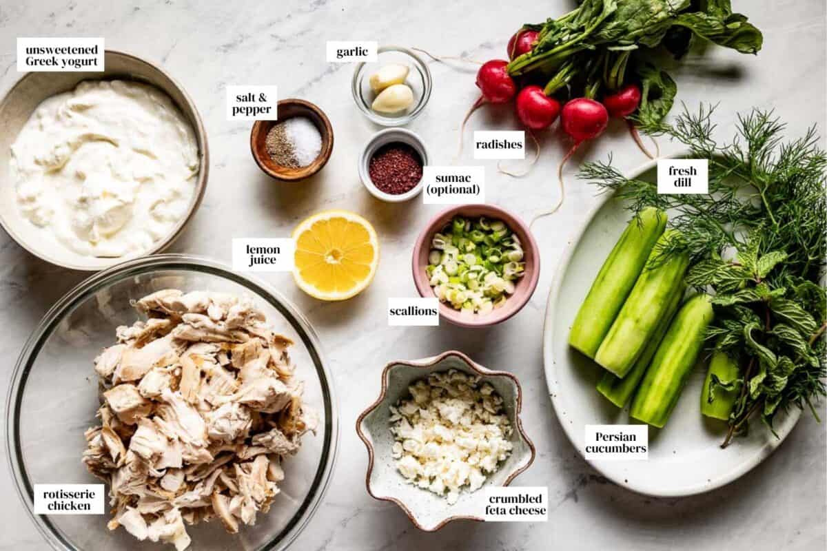 A flat-lay of labeled ingredients for Mediterranean chicken salad including rotisserie chicken, peeled Persian cucumbers, Greek yogurt, and fresh radishes.