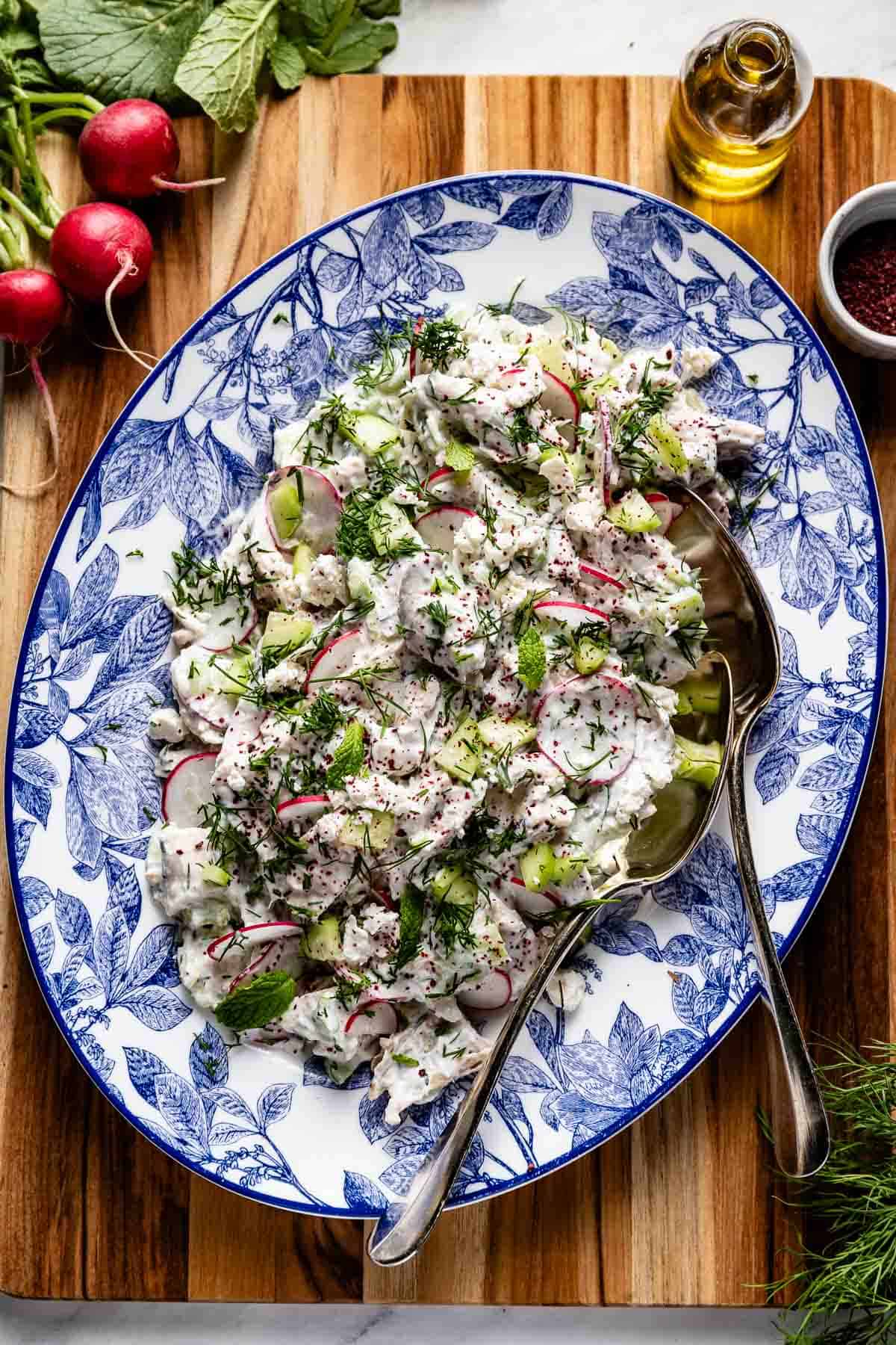 A portrait view of the final Tzatziki Chicken Salad on a decorative blue and white patterned platter with peeled diced cucumbers, radishes, feta, fresh dill, and sumac, on a wooden board.