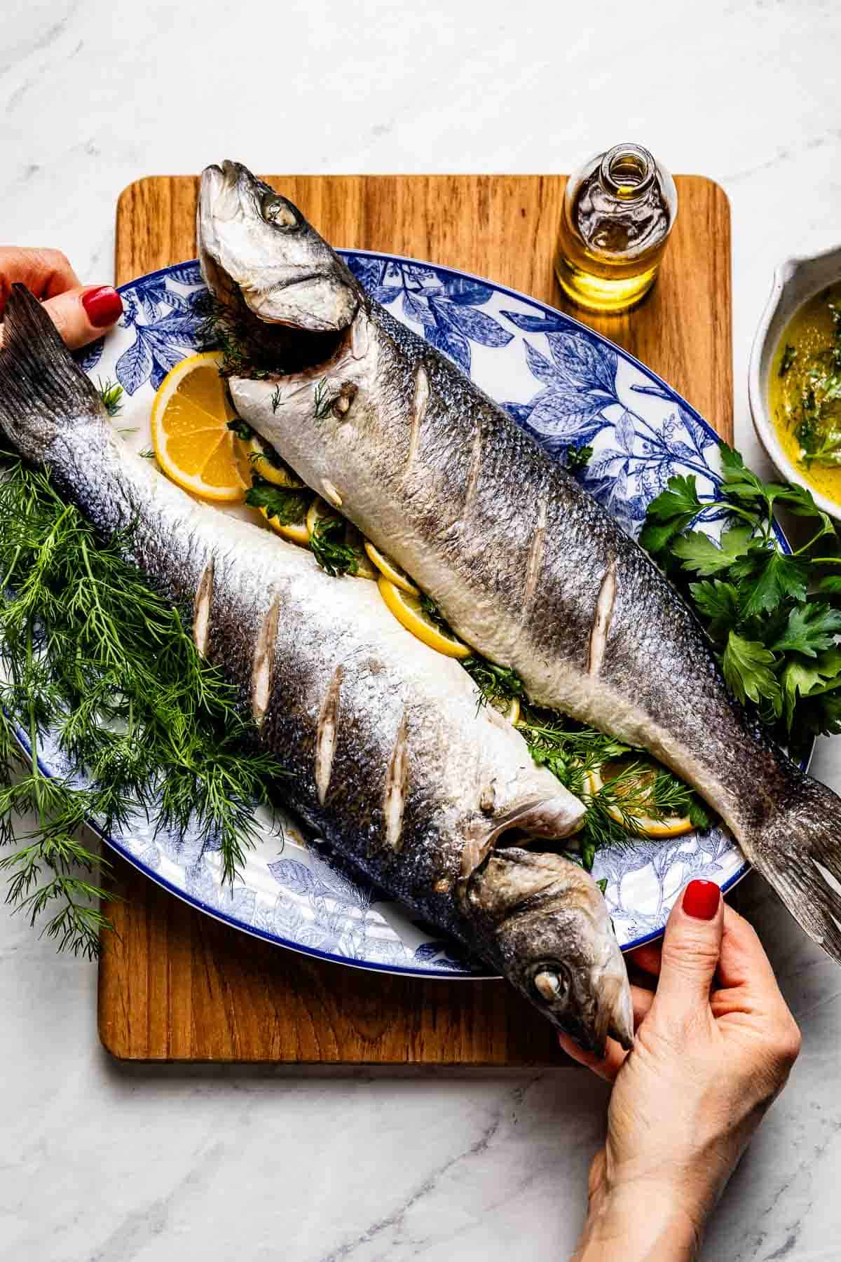 A woman's hand presenting a platter of two whole roasted branzino Mediterranean style, garnished with fresh herbs and lemon slices with lemon olive oil sauce on the side.