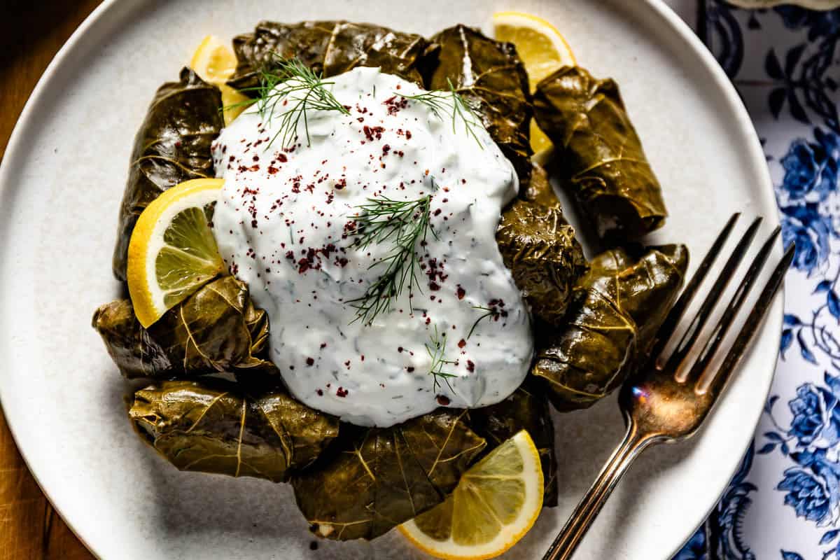 A plated serving of Mediterranean stuffed grape leaves piled on a white plate with fresh lemon wedges, a bowl of dill yogurt sauce on the side, and sumac in a small dish.