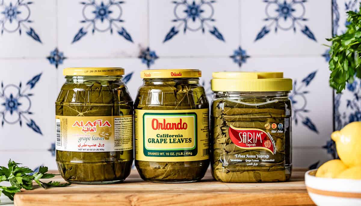 Three brands of jarred grape leaves in brine (Alafia, Orlando California, and Sarim) displayed on a wooden board.