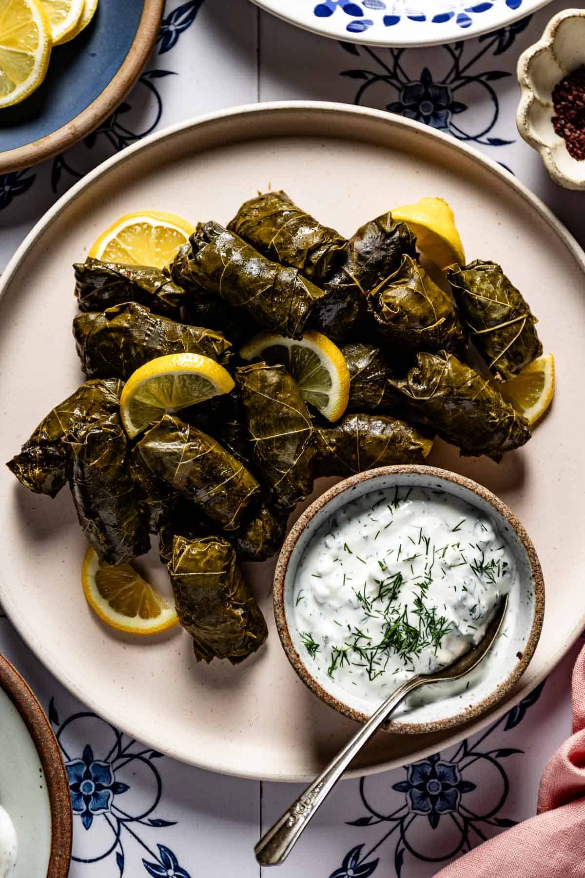 Stuffed grape leaves piled on a plate, garnished with lemon slices and served with a bowl of yogurt dill sauce.